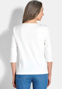 Weißes Langarmoberteil mit quadratischem Ausschnitt. Glatte Textur und lockere Passform, kombiniert mit blauen Jeans mit gestickten Taschendetails.