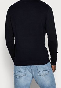 Mörkblå långärmad tröja med ribbad halsringning och ärmslut, tillsammans med ljusblå denimjeans med vinklade bakfickssömmar.