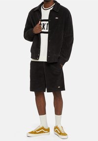 Czarna kurtka sztruksowa z zatrzaskami, biała sweter z czarnym paskowanym wzorem, czarne szorty i musztardowe trampki z białymi akcentami.