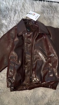 Giacca in similpelle marrone lucida con colletto classico, chiusura con zip e due tasche frontali. Presenta un orlo inferiore arricciato. Etichetta inclusa.