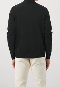 Mann in einem schwarzen, langärmeligen Rollkragenpullover und cremefarbenen Jeans, von hinten vor einem schlichten Hintergrund gezeigt.