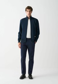 Jūras spēku zip-up jakas ar augstu apkakli, apvienotas ar baltu t-kreklu un tumšām biksēm. Melni uzvalka apavi noslēdz tēlu.