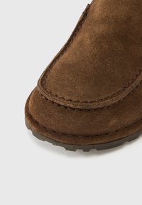 Καφέ σουέτ παπούτσι χωρίς κορδόνια με στρογγυλεμένη μύτη, λεπτομερείς ραφές κατά μήκος της άκρης και υφή στη μαύρη καουτσούκ σόλα.