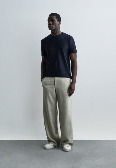 Άνδρας στέκεται με τα χέρια στις τσέπες, φορώντας μπλε κοντομάνικο πουκάμισο, μπεζ παντελόνι φαρδύ και λευκά αθλητικά παπούτσια σε γκρι πάτωμα.