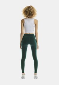 Mujer con cabello rizado de pie dando la espalda, vistiendo un top corto blanco, leggings verde oscuro y zapatillas deportivas blancas sobre un fondo sencillo.
