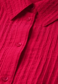 Camicia rossa brillante con colletto, realizzata in un tessuto testurizzato, con patta di bottoni frontale e dettagli superficiali testurizzati e stropicciati.