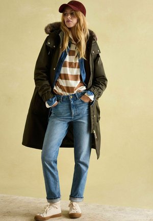 Parka jacket in dark green with a fur-lined hood, layered over a striped brown and white shirt, denim jacket, and blue jeans. Cream sneakers.