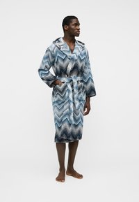 Uomo che indossa un accappatoio blu e bianco con cappuccio a motivo a chevron, in piedi a piedi nudi su uno sfondo chiaro e uniforme.