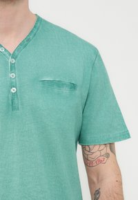 Άνδρας με πράσινο κοντομάνικο πουκάμισο Henley με κουμπιά και τατουάζ τριαντάφυλλου που δείχνει μια ημερομηνία στο εσωτερικό του δεξιού πήχη.