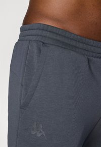 Mørkegrå joggebukser laget av mykt stoff har en teksturert midjebånd, sidelommer og en subtil logo på låret.