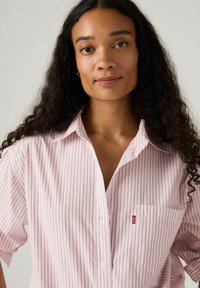 Camicia rosa chiaro a maniche corte con righe verticali bianche e rosse, chiusura con bottoni davanti e taschino sul lato sinistro con un piccolo logo.