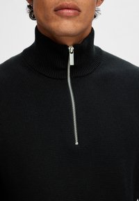 Black knit turtleneck with a silver zipper, ribbed texture, and a fitted design. Close-up view focuses on the neckline and zipper details.