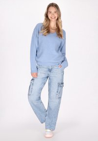 Pulover bleu deschis cu guler în V, croială lejeră, asortat cu blugi cargo de nuanță deschisă, cu buzunare laterale. Modelul poartă pantofi sport pastel.