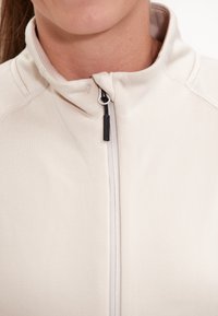 Ljus beige zip-up jacka med ståkrage, slätt tyg och kontrasterande dragkedjedetalj. Sömmarna ger struktur åt designen.