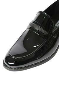 Mocassino in pelle nera lucida con una cintura decorativa e dettagli di cucitura su uno sfondo bianco, evidenziando la finitura lucida della scarpa.