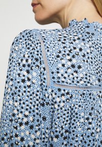Blouse bleu clair avec motif d'étoiles en noir et blanc. Présente un col à volants et des coutures détaillées le long du yoke. Tissu doux et léger.