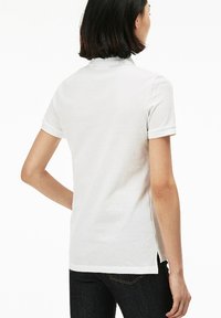 Weißes Polo-Shirt mit kurzen Ärmeln, strukturiertem Stoff, geripptem Kragen und seitlichem Schlitz. Rückansicht dargestellt, mit dunkler Jeans, die am unteren Ende sichtbar ist.