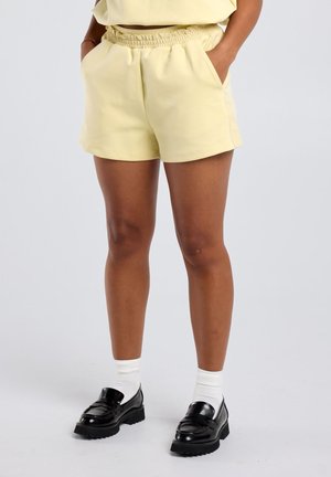 Persoon met handen in de zakken, draagt bleke gele shorts met elastische taille, witte sportsokken en zwarte leren loafers op een grijze achtergrond.