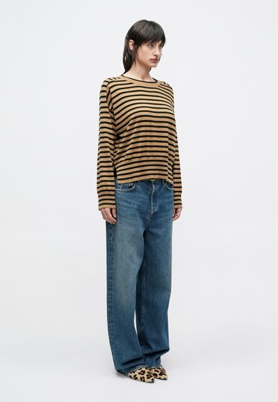 Ριγέ μακρυμάνικη μπλούζα σε καφέ και μαύρες απ οχρώσεις, συνδυασμένη με φαρδιά μπλε τζιν και παπούτσια με ζωικό print, σε απλό λευκό φόντο.