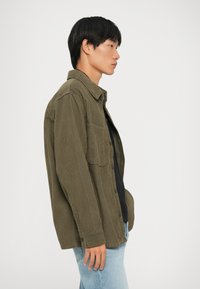 Camicia in velluto a coste verde oliva con bottoni sul fronte, taschino sul petto e maniche lunghe, indossata sopra una maglietta nera e abbinata a jeans chiari.