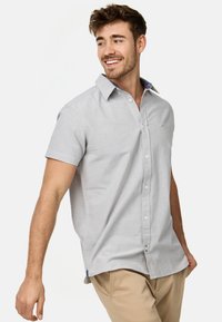 Camisa de manga corta gris claro con cuello botón. Presenta un sutil detalle de rayas en el cuello y un pequeño logo en el pecho.