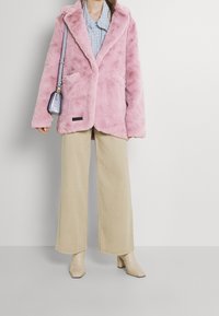 Roze nepbont jas met een sjawlboord, voorzakken, gecombineerd met beigekleurige wijde broek en lichte haklaarzen. Kleine schoudertas.