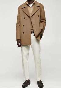Abrigo de lana marrón de doble botonadura sobre un suéter beige con cremallera, combinado con pantalones crema y zapatos oscuros de tipo slip-on, de pie contra un fondo claro.
