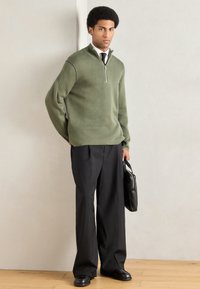 Πράσινο πλεκτό πουλόβερ με φερμουάρ και ψηλό γιακά, συνδυασμένο με μαύρο παντελόνι με φαρδιά πόδια και μαύρα παπούτσια, κρατώντας μια μαύρη τσάντα ώμου.