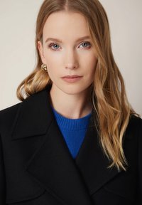 Czarny wełniany płaszcz z dużymi klapami noszony na niebieskim swetrze dzianinowym. Posiada falowane włosy do ramion oraz okrągłe złote kolczyki. Neutralne tło.