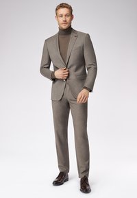 Szary klasyczny garnitur o slim fit, jedno zapięcie, klapy typu notch oraz spodnie. Pod spodem brązowy golf. Brązowe eleganckie buty.