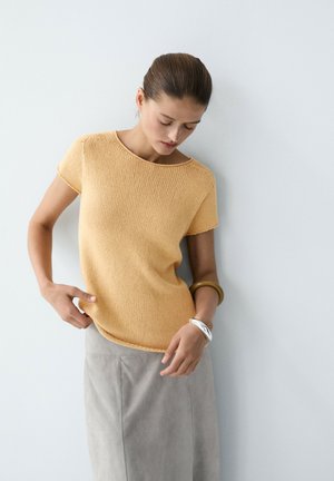 Femme portant un haut en maille jaune à manches courtes et une jupe gris clair, debout contre un mur blanc, regardant vers le bas, les mains près de la taille.
