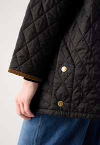 Quilted black jacket with a textured surface, featuring gold buttons and a contrasting brown trim at the cuffs. Blue denim visible below.