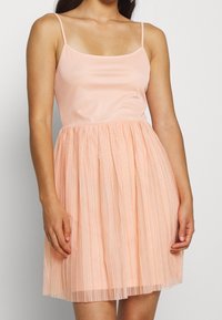 Femme portant une robe pêche sans manches avec un corsage ajusté et une jupe plissée, debout devant un fond blanc uni.