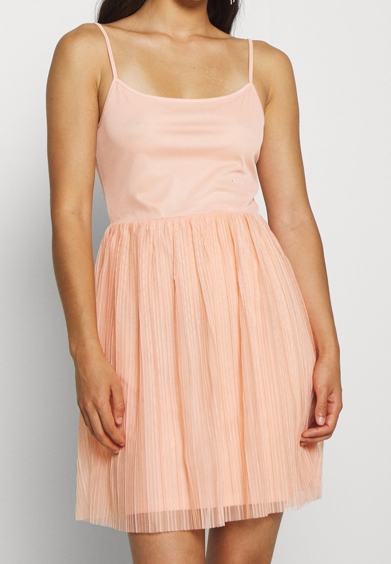 Femme portant une robe pêche sans manches avec un corsage ajusté et une jupe plissée, debout devant un fond blanc uni.