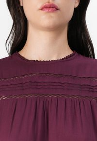 Vinröd blus med veckad design, texturerade spetsinsatser och en scalloped halsringning. Slätt tyg med en flödande silhuett.