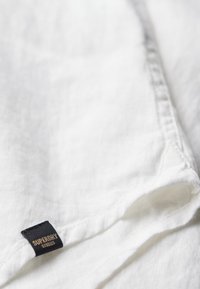 Biała lniana koszula o teksturowanym wykończeniu, z markową czarną metką z złotym napisem u dołu. Naturalny, miękki materiał.