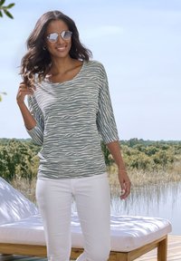 Leichtes, langärmliges Oberteil im grünen und weißen Zebramuster, kombiniert mit weißen Jeans. Das Modell trägt eine Sonnenbrille und steht im Freien.