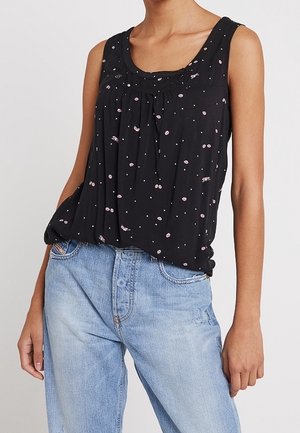 Woman wearing sleeveless black blouse with small pink floral patterns and light blue high-waisted jeans, standing against a plain background.