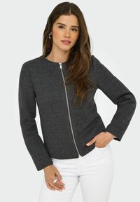 Veste à motif chevron noir avec des manches longues, une fermeture éclair à l'avant et un col rond. Portée sur un pantalon blanc.