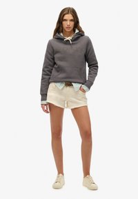 Un sweat à capuche gris avec un logo "Superdry" texturé, un short crème avec un ourlet effiloché, et des baskets beige. Une chemise à col bleu clair sous le sweat.