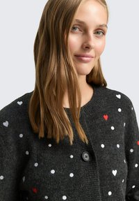 Grauer Strickcardigan mit weißen und roten Herzmustern sowie Punkten, ausgestattet mit einem Rundhalsausschnitt und schwarzen Knopfverschlüssen. Weiche Textur.