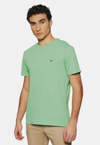 T-shirt vert clair à col rond fabriqué en coton doux. Il présente des manches courtes et un petit logo brodé sur la poitrine gauche.