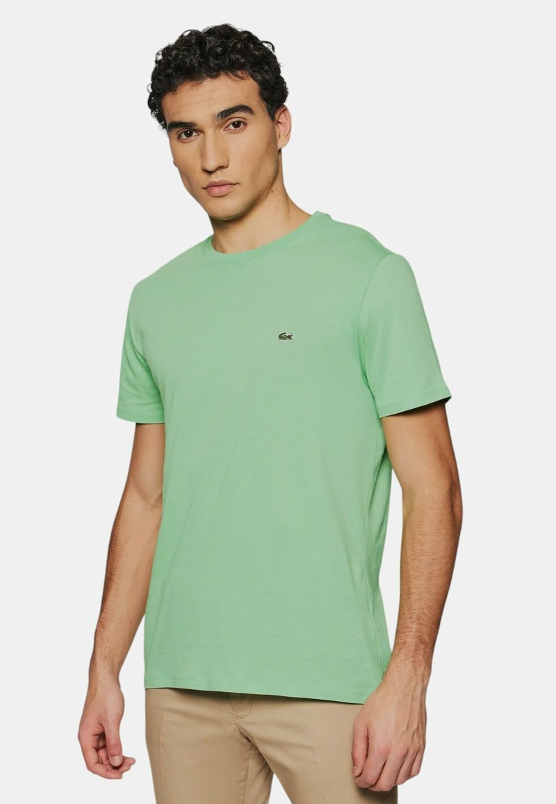 T-shirt vert clair à col rond fabriqué en coton doux. Il présente des manches courtes et un petit logo brodé sur la poitrine gauche.