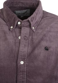 Corduroy shirt in dark purple with vertical ridges, button-down collar, and black logo. Features white plastic buttons and a pocket detail.