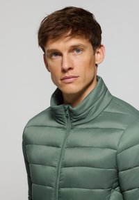 Chaqueta acolchada verde con cuello alto y cremallera frontal. La tela es suave con patrones de costura horizontal para el aislamiento.