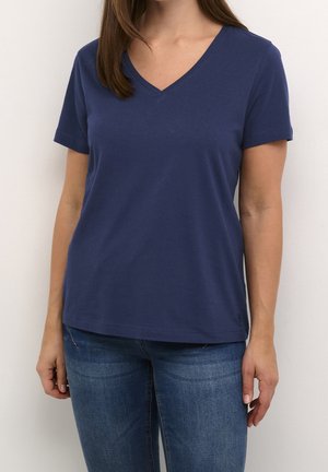 Nainen pukeutuneena yksinkertaiseen tummansiniseen, lyhythihaiseen V-kauluspaitaan ja sinisiin farkkuihin, seisoo yksivärisen vaalean taustan edessä.