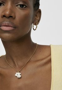 Boucle d'oreille en or et un collier en perles sur un cou avec une peau lisse et uniforme, portant un haut à rayures jaune clair. Gros plan sur les accessoires.
