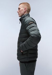 Vadderad jacka i mörkgrått, med hög krage, dragkedjefickor och en texturerad, quiltad finish med en logo på ärmen.