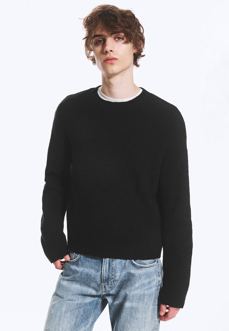 Jeune homme aux cheveux bouclés portant un pull en maille noir par-dessus une chemise blanche et un jean bleu clair, debout devant un fond blanc uni.