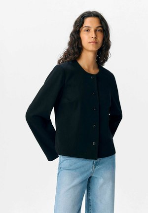 Giacca nera corta con scollo rotondo, maniche lunghe e chiusura frontale con bottoni. Texture liscia, design minimal, abbinata a jeans blu chiaro.